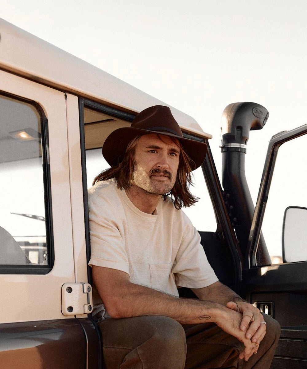 Packable Felt Fedora Male model leaning on an off-road vehicle wearing the Indy premium brown felt fedora for a rugged look